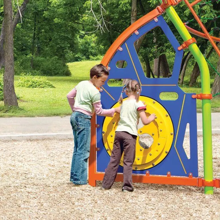 Two children interact with a colorful play panel featuring a wheel and geometric shapes on a sunny day.