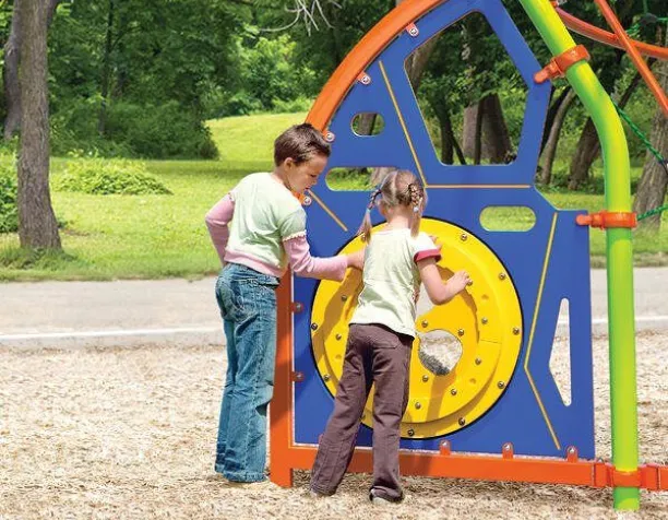 Two children interact with a colorful play panel featuring a wheel and geometric shapes on a sunny day.