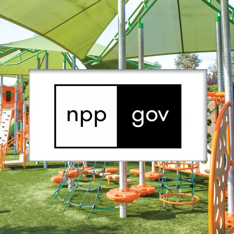 Green and orange playground with rope net and climbing panels under large shade canopy.