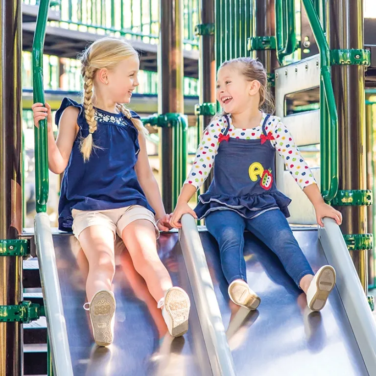 Two young girls smiling and sliding down parallel metal slides in a playground.