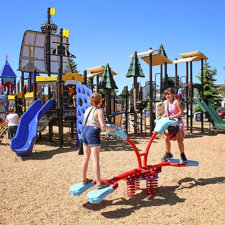 Two children play on a blue and red spring rider in a playground under a clear blue sky.