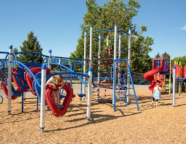 Children playing on blue and red climbing structures and slides on a sunny day with wood chip surfacing.