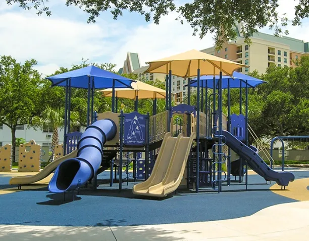 Playground with blue and beige slides, climbers, and shade structures under trees and near buildings.