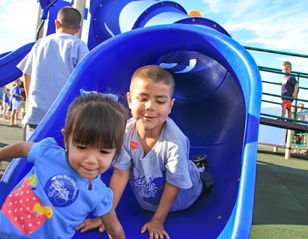 Children sliding down a bright blue tunnel slide on a sunny day.