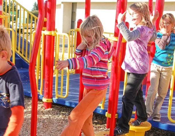 Children walking on a balance beam in bright sunlight, surrounded by red and yellow playground bars.
