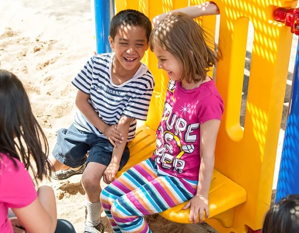 Two children laughing and sitting on a yellow playground panel with sand below.