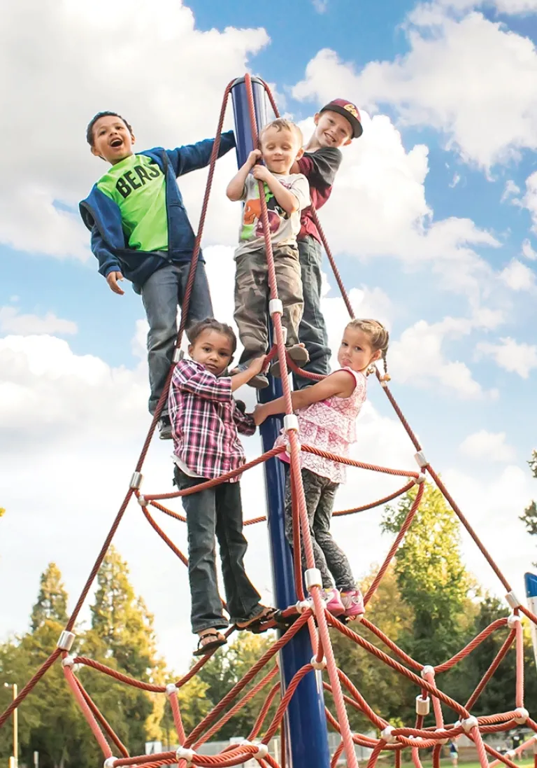 Five children climbing a red rope pyramid under a blue sky.