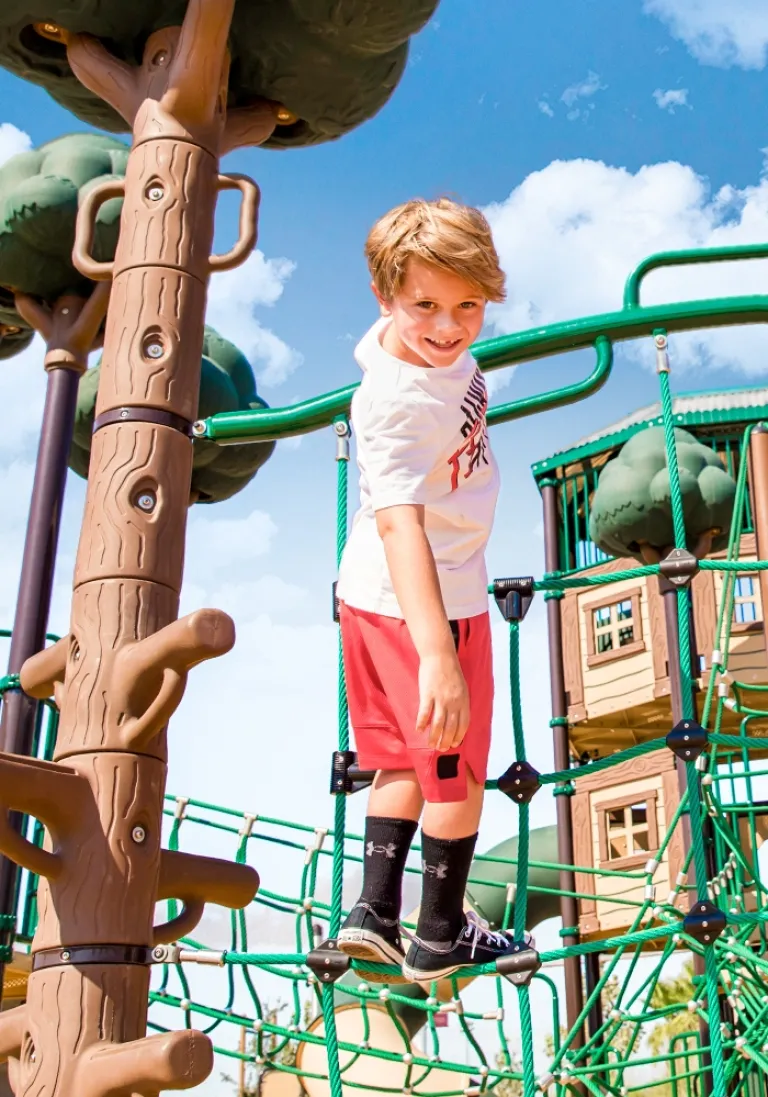 Smiling child walking on a rope bridge next to tree-like structures under a blue sky.