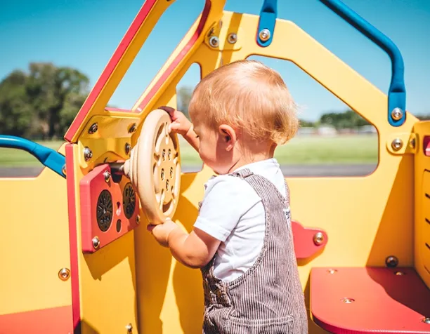 Toddler plays with steering wheel on bright yellow and red playground structure.