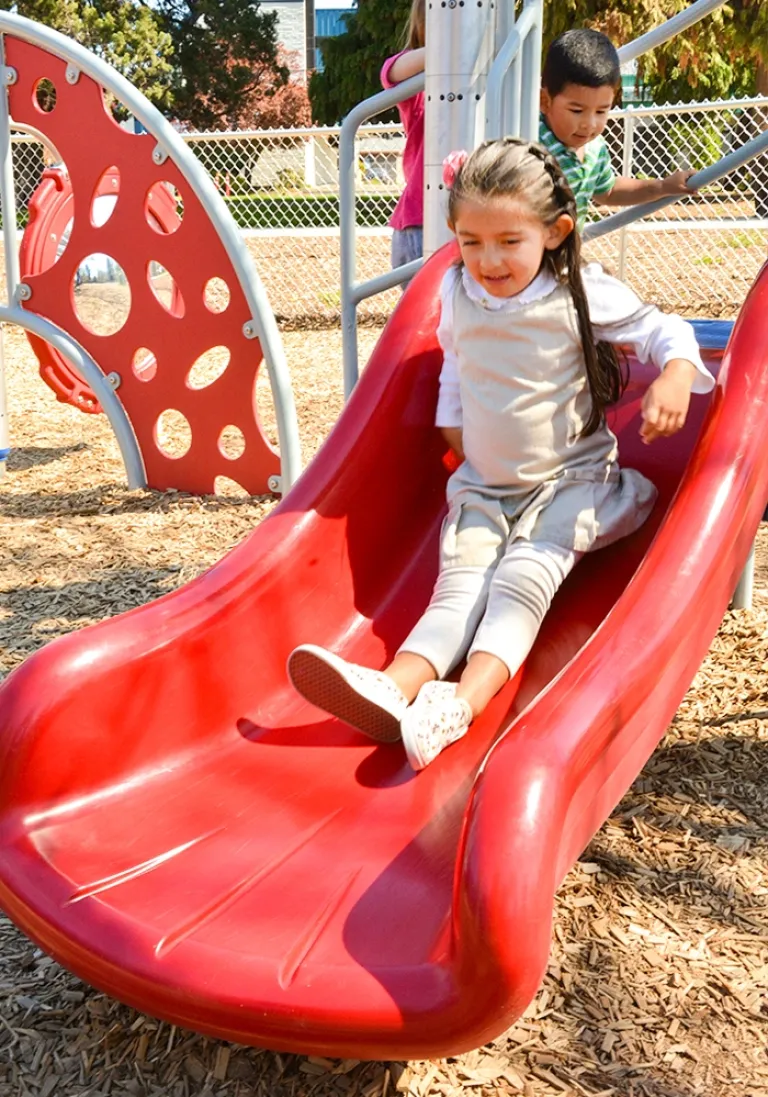 Girl sliding down a red slide with a boy climbing in the background on a sunny day.
