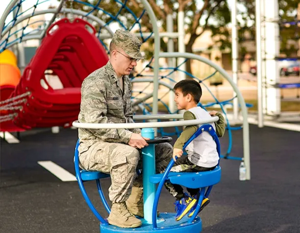 Person in military uniform and child sitting on a blue playground spinner.