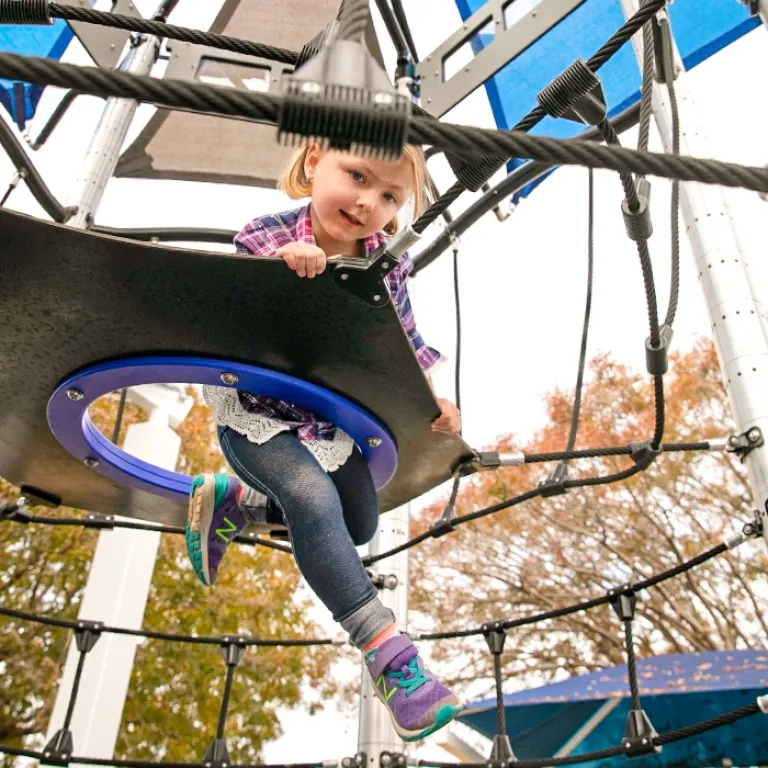 Child climbing through a circular opening in a rope net structure, wearing colorful shoes and surrounded by autumn trees.