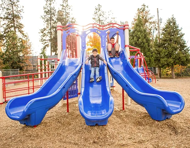 Two children on a triple blue slide with woodchip surfacing, surrounded by trees and sunlight.