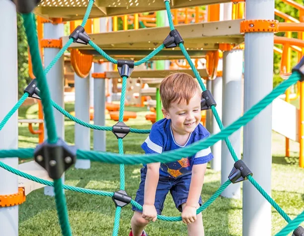 Smiling toddler climbing on a green rope net structure with orange and gray playground equipment in the background.