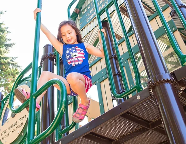 Girl in patriotic outfit slides down green fireman's pole on playground.
