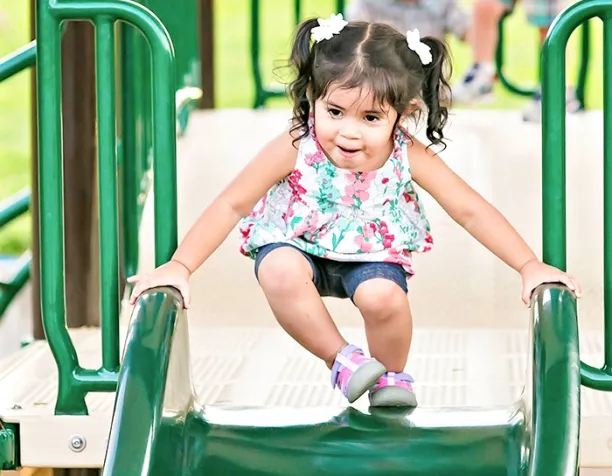 Toddler in a floral shirt sliding down a green slide at the playground.
