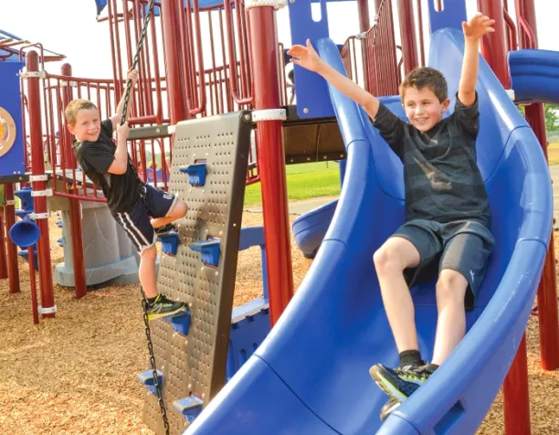 Two boys enjoy a blue slide and climb on a wall panel at a playground with wood chip surfacing.