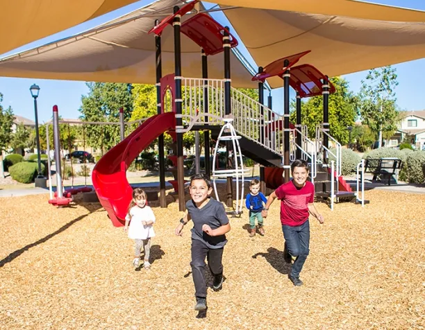 Four children running in front of a playground structure with slides and shade canopies on a sunny day.
