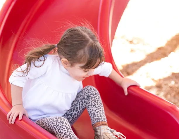 Young girl in white shirt sliding down a red slide on a sunny day.