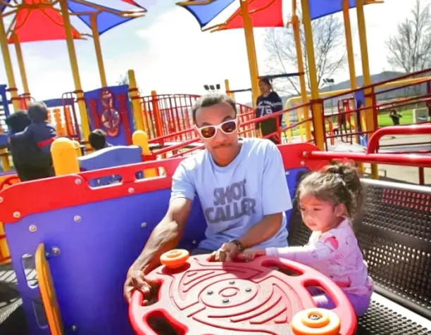 Adult and child interacting with a red and orange steering panel under a shade structure on a sunny day.