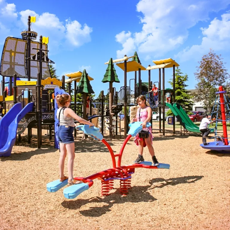 Two girls balancing on a red spring seesaw in a playground with slides and climbing structures.