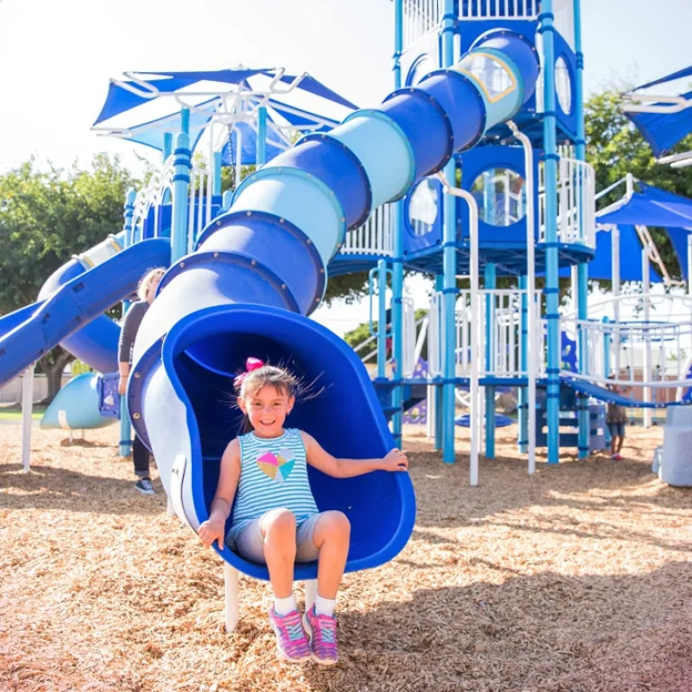 Child in a striped dress sliding down a blue spiral slide on a sunny day.