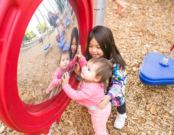 Two children interacting with a red circular mirror in a playground area with wood chips.