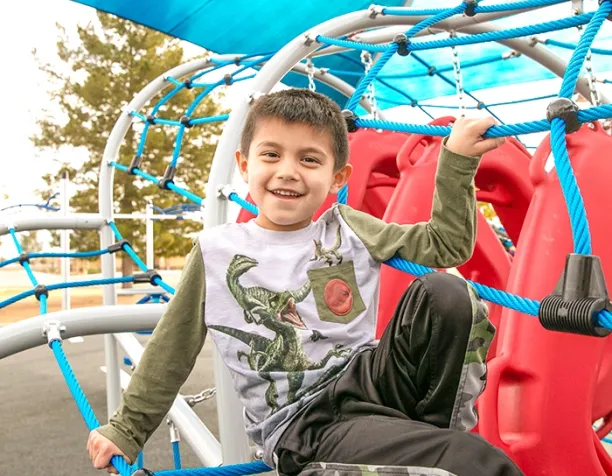 Smiling boy sits on a climbing structure with blue ropes and a red slide.