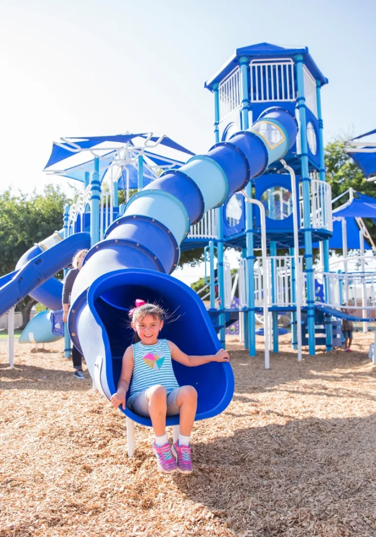 Smiling girl in a striped dress sliding down a blue spiral slide on a sunny day.
