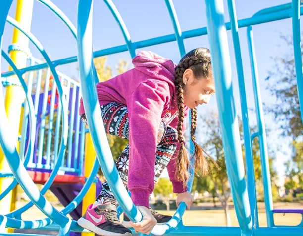 Girl in a pink hoodie climbing a blue circular play structure on a sunny day.
