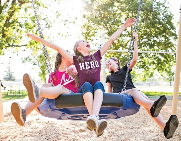 Three children joyfully swinging on a round blue group swing under sunny skies.