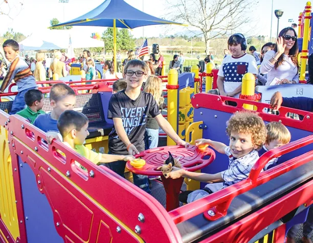 Children interact on a colorful play structure with red panels and a spinning element on a sunny day.