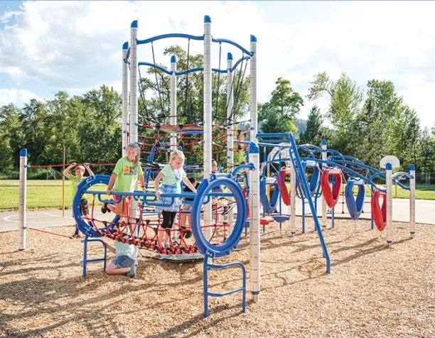 Children climb and stand on a blue and red rope structure under a sunny sky with wood chip surfacing.