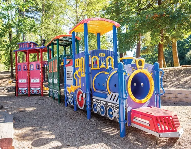 Colorful train-themed play structure with blue, yellow, red, and green sections under tall trees.
