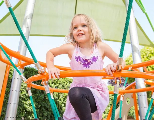 Smiling child climbing on orange rope structure under a green canopy.