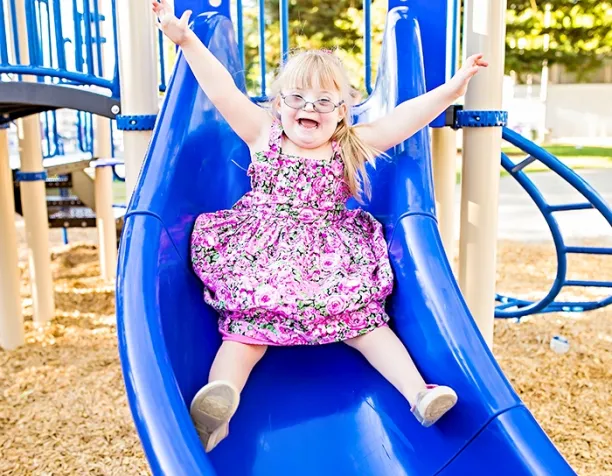 Smiling girl with glasses sliding down a blue slide wearing a floral dress on a sunny day.