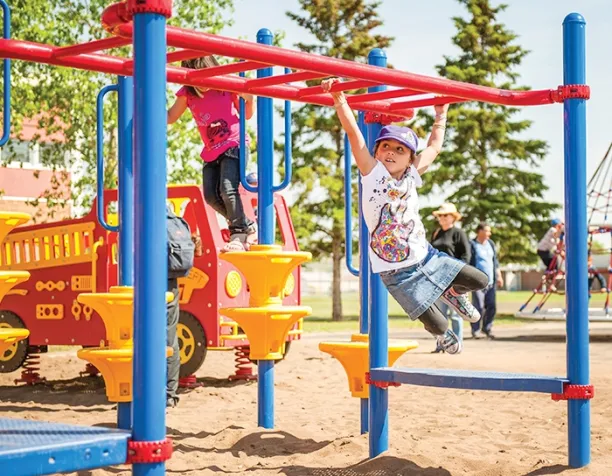 Child swinging from red monkey bars near a fire truck play structure under a bright sky.