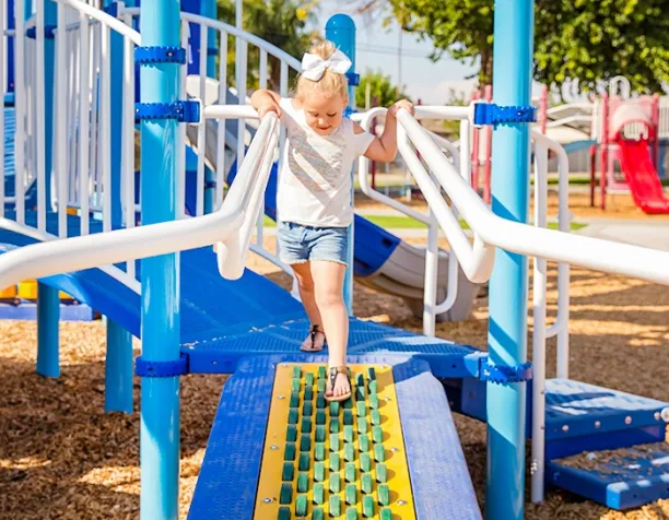 Young girl walking across a textured bridge on a playground with blue and white structures.