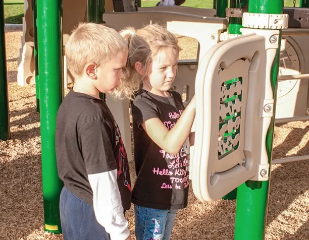 Two children interact with a play panel on a sunny playground with wood chip surfacing.