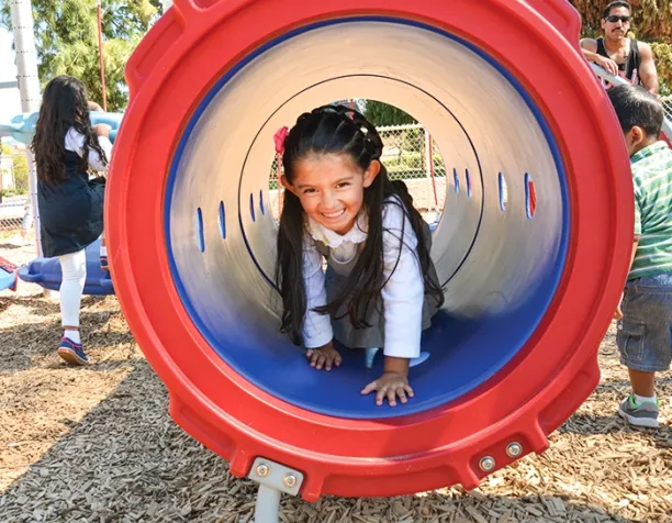 Young girl crawling through a red tunnel tube on a sunny day.