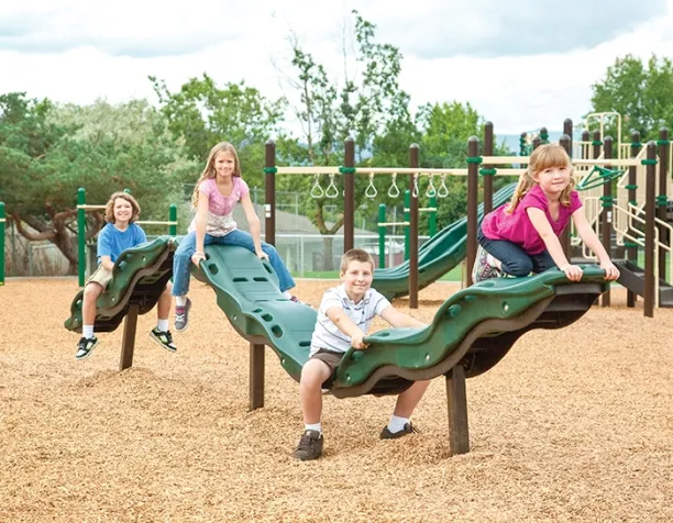 Four children smiling and balancing on wavy green beams in a playground on a cloudy day.