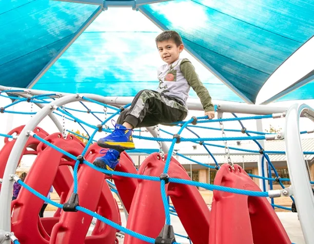 Child climbing on a rope net structure with red panels under a blue shade canopy.