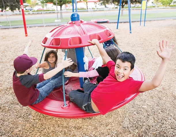 Four children spin joyfully on a red merry-go-round on wood chips.