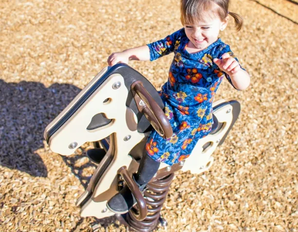 Smiling girl in a floral dress rides a spring animal toy on wood chip surfacing.