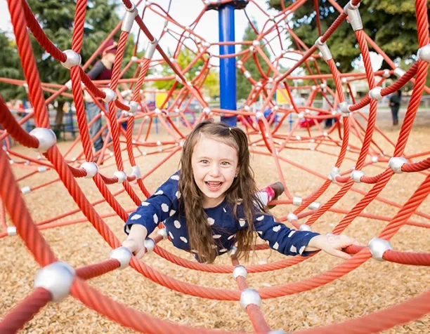 Smiling girl crawling on a red rope net structure with wood chip surfacing in a park.