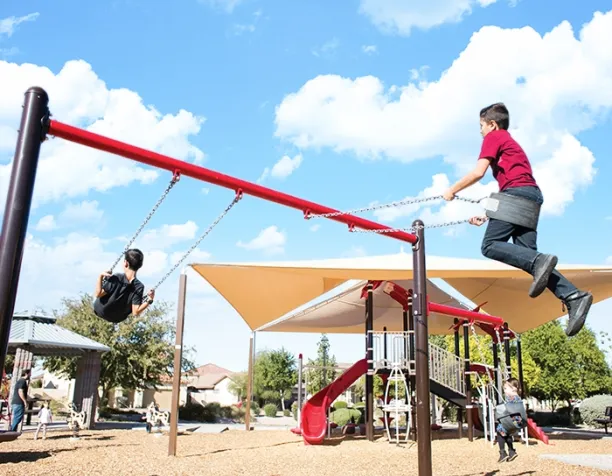 Two boys swinging on swings under a clear blue sky with shade structures nearby.