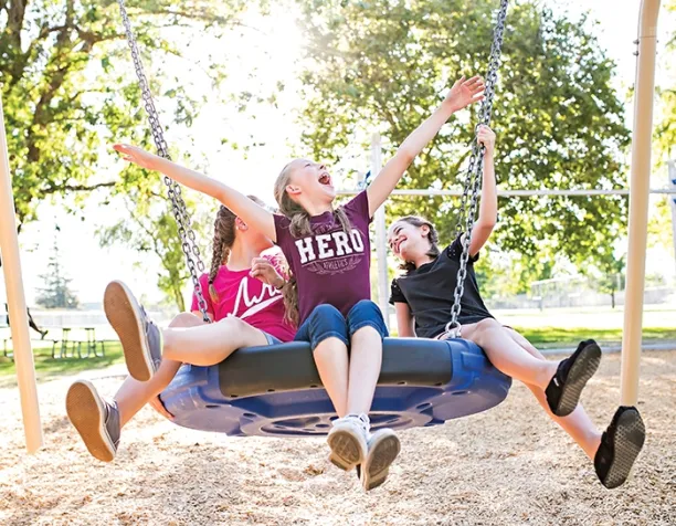 Three kids joyfully swinging on a blue disc swing, with wood chip surfacing and trees in the background.