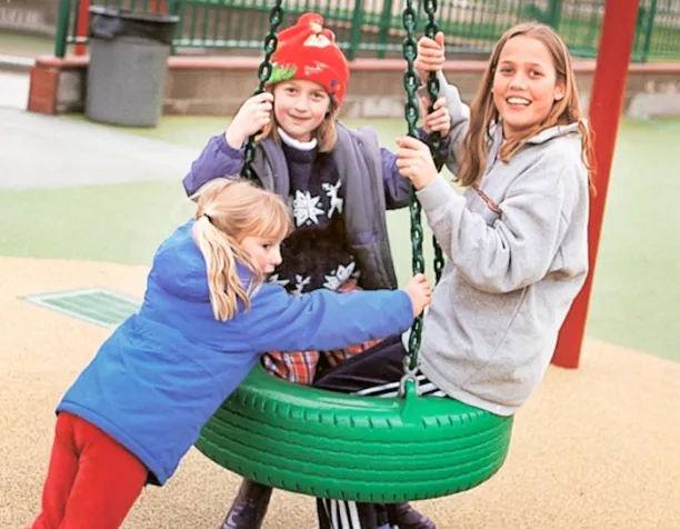 Three children smiling and playing on a green tire swing at a playground.