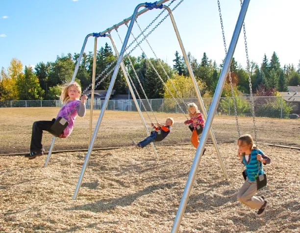 Four children swing on metal swings under a clear blue sky surrounded by trees.