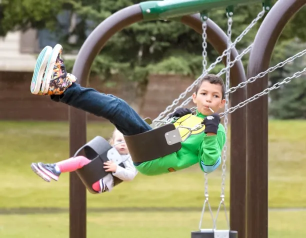 Two children swinging on belt swings, one wearing a green shirt, in a grassy area with trees in the background.
